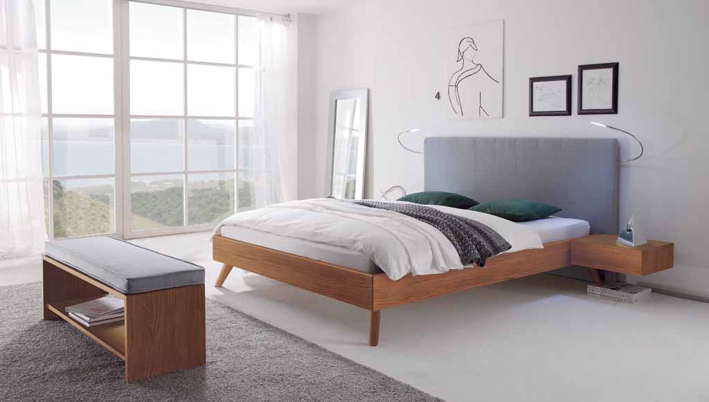 Hasena Wood-Line Bettanlage | Eiche brandy, geölt | Wandpaneel Stoff grau | Leselampe Helles Schlafzimmer mit einem großen, modernen Bett aus Holz und einem grauen Kopfteil, flankiert von Nachttischen, vor großen Fenstern mit Blick auf eine malerische Landschaft.