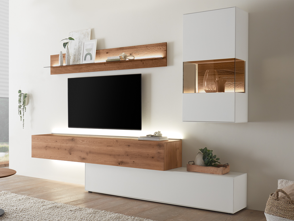 Eine moderne Wohnwand von Hartmann Torido in einem hellen Wohnzimmer. Die Wohnwand besteht aus einem schwebenden TV-Element aus Holz, einem hohen weißen Schrank mit beleuchtetem Glasfach und einem beleuchteten Wandregal darüber.