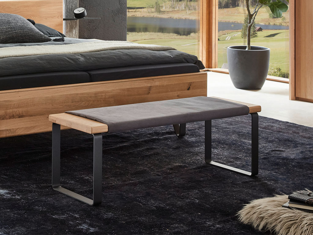 Braune Bank mit grauem Sitzkissen und schwarzen Metallkufen vor einem Bett in einem stilvoll dekorierten modernen Raum