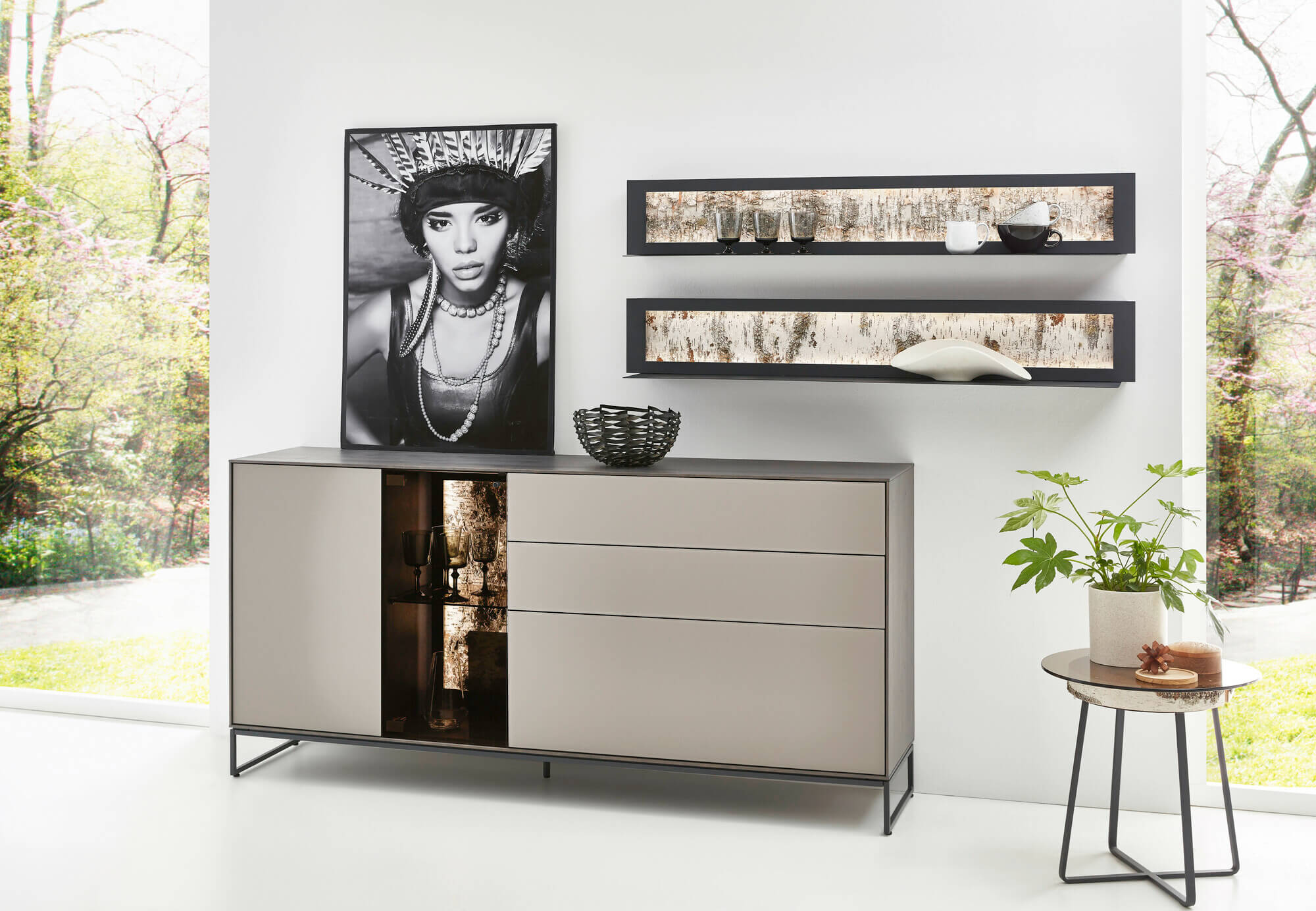 Sideboard mit beleuchtetem Vitrinenfach und grauer Front. Darüber hängen zwei Designer Regalböden.