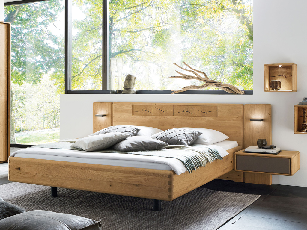Wöstmann WSM 1600 - Bettanlage | Wildeiche massiv | konfigurierbar Ein stilvoll eingerichtetes Schlafzimmer mit einem modernen Holzbett, dekorativen Kissen und einem großen Fenster mit Ausblick ins Grüne