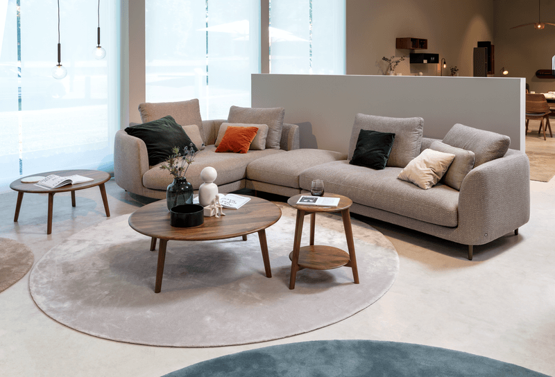 Ein stilvoll eingerichtetes Wohnzimmer mit einem grauen Ecksofa, farbigen Kissen und runden Holztischen auf einem Teppich vor großen Fenstern.