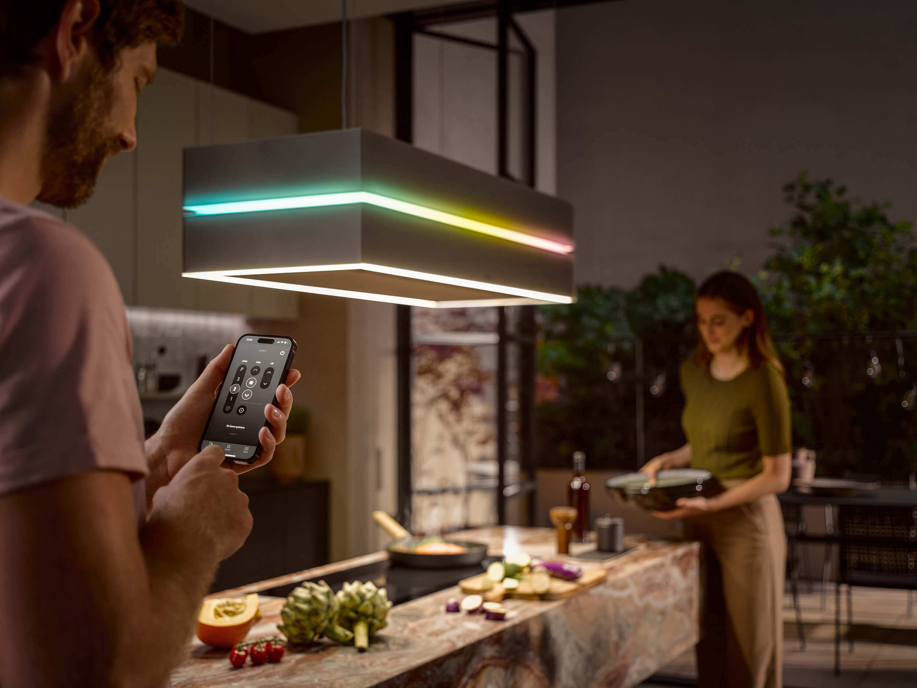 Ein Mann bedient eine Dunstabzugshaube über eine Smartphone-App, deren Licht in mehreren Farben leuchtet. Im Hintergrund bereitet eine Frau Essen auf einer Kücheninsel mit einer unverwechselbaren rotbraunen Marmorplatte zu.