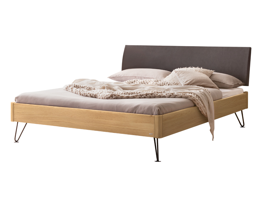 Freigestelltes Bett mit einem Bettrahmen aus heller Eiche, einem anthrazitfarbenen Kopfteil und schwarzen, schmalen Haarnadelbeinen. Das Bett ist mit hellgrauer Bettwäsche und einem cremefarbenen Strickplaid dekoriert.