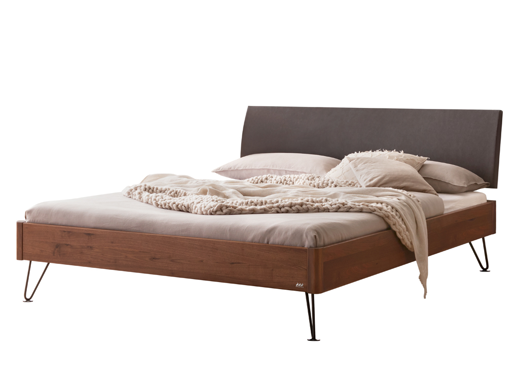  Freigestelltes Bett mit einem Bettrahmen aus dunklem Nussbaumholz, einem anthrazitfarbenen Kopfteil und schwarzen, schmalen Haarnadelbeinen. Das Bett ist mit hellgrauer Bettwäsche und einem cremefarbenen Strickplaid dekoriert.