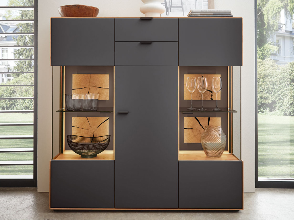 Wöstmann WM 2410 - Highboard | Type 4340 | Lack und Wildeiche massiv | Hirnholzakzent Moderner grauer Wohnzimmerschrank mit integrierten Glasregalen, gefüllt mit Gläsern und dekorativen Elementen, steht vor einem Fenster.