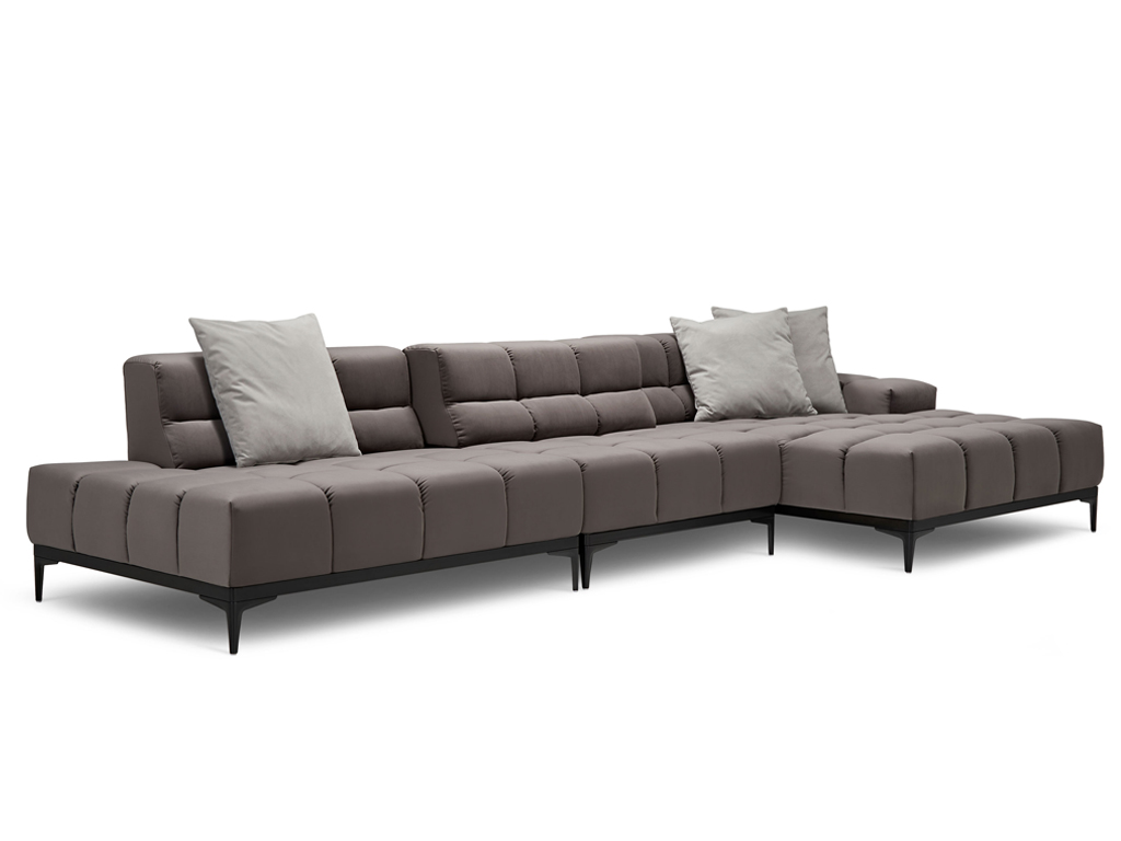 Ein langes L-förmiges Sofa in einem dunklen Taupe-Ton mit gesteppter Sitzfläche und Rückenlehne. Es steht auf schlanken schwarzen Metallfüßen. Zwei helle, graue Zierkissen liegen auf der Rückenlehne. Der Hintergrund ist weiß.