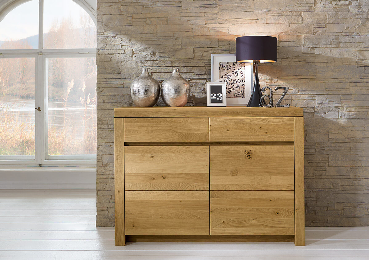 Wöstmann Soleo 3000 - Sideboard | Type 2350 | B: 131,2 cm | konfigurierbar Eine stilvolle Eichenkommode vor einer Steinwand, dekoriert mit zwei Silbervasen, einem Bild, einer Tischlampe und einem Kalenderblock. Ein großes Fenster lässt Tageslicht in den Raum.