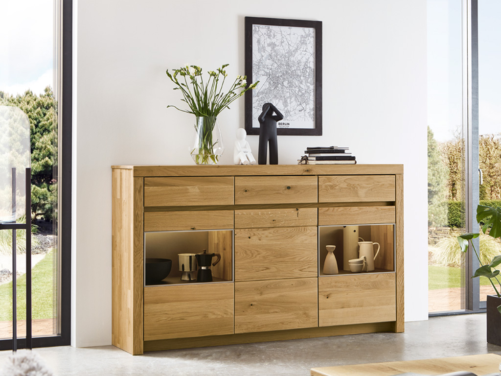 Wöstmann Soleo 3000 - Highboard | Type 3940 | B: 190,1 cm | konfigurierbar Helle Holzkommode mit Glasfenstern, geschmückt mit Pflanzen und Keramikfiguren, in einem modernen Wohnzimmer mit großen Fenstern.