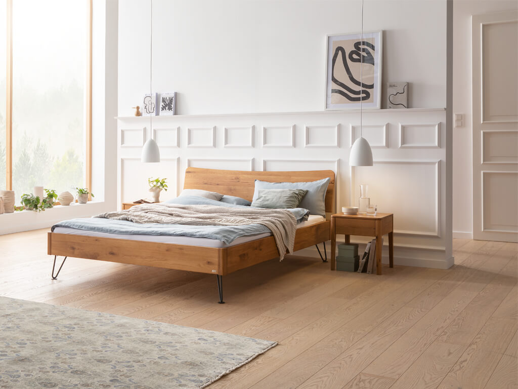 Ein Hasena Bett Locarno in Wildeiche natur geölt, in einem hellen Schlafzimmer mit minimalistischer Dekoration. Die schwarzen, anthrazitfarbenen Haarnadel-Beine sind sichtbar. Auf der linken und rechten Seite stehen passende Nachttische.