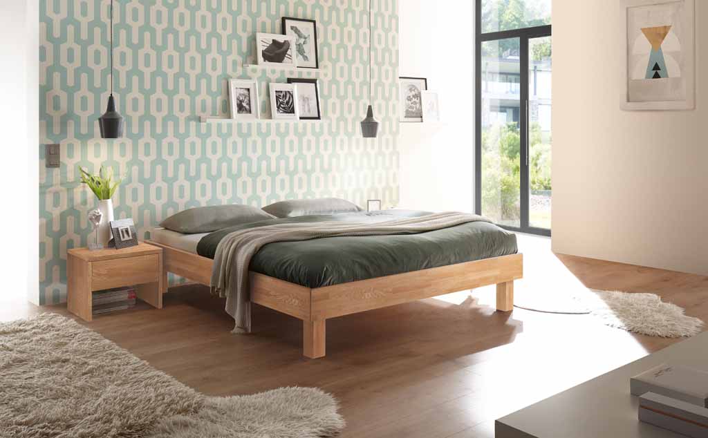 Helles Schlafzimmer mit einem Holzbett, grüner Bettwäsche, Nachttisch mit Dekoration, geometrischer Tapete und großem Fenster.