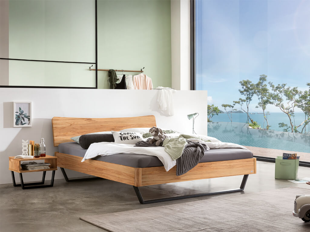 Ein Hasena Bettmodell Tresali aus Wildbuche, das in einem hellen, modern eingerichteten Schlafzimmer mit großen Fenstern und Blick auf den Pool steht.