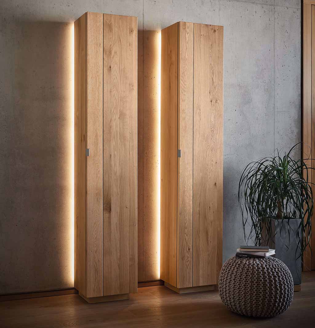 Wimmer Signatura | Zeilenschrank mit Beleuchtung | 154110/154111 Zwei hohe, schmale Holzschränke mit integrierter LED-Beleuchtung, die eine warme Atmosphäre vor einer Betonwand schaffen, daneben ein geflochtener Pouf.