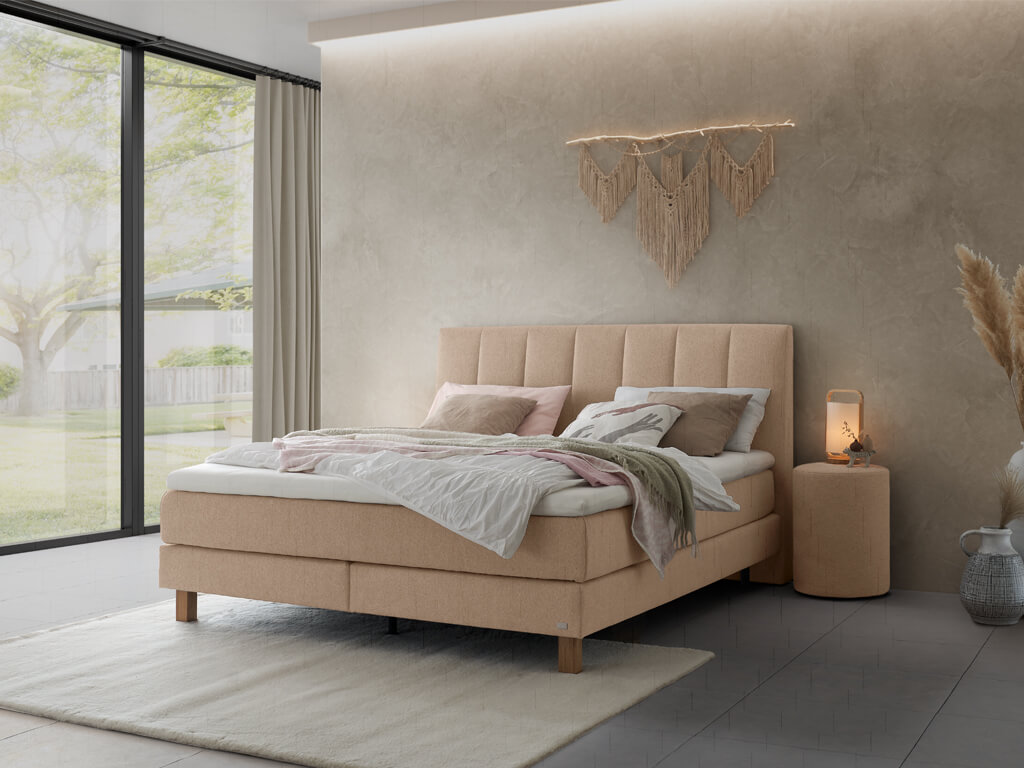 Ein helles Schlafzimmer mit einem Boxspringbett in einem warmen Beigeton und einem großen Fenster, das eine ruhige Umgebung zeigt.