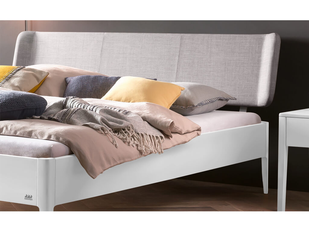 Eine Nahaufnahme des oberen Teils eines Bettes Pienza in Weiß-deckend-lackiert. Das Bett hat ein gepolstertes Kopfteil in hellem 'Pepe-grey' Stoff und ist mit verschiedenen Kissen in Beige, Grau und Gelb sowie einer gestrickten Decke dekoriert.