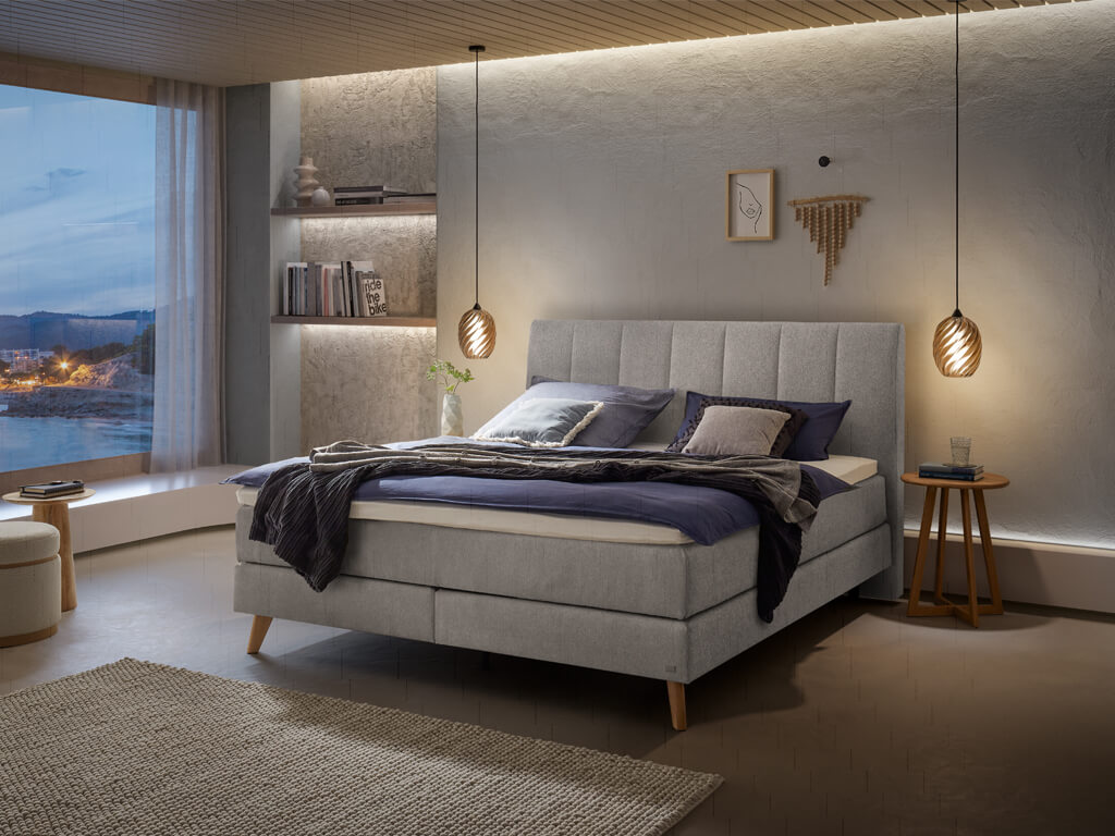 Ein Boxspringbett in hellgrauer Stoffpolsterung und Holzfüßen in einem modern gestalteten Schlafzimmer bei Nacht.