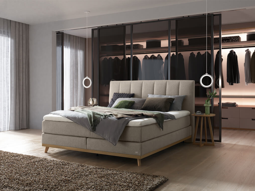 Ein Hasena Boxspringbett Pronto Stefania Roy mit einem hellgrauen Stoffbezug, das stilvoll vor einem eleganten begehbaren Kleiderschrank präsentiert wird.