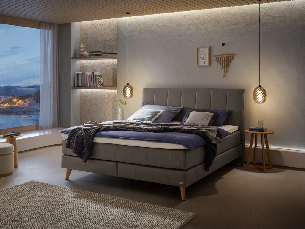 Ein Boxspringbett in dunkelgrauer Stoffpolsterung und Holzfüßen in einem modern gestalteten Schlafzimmer bei Nacht.