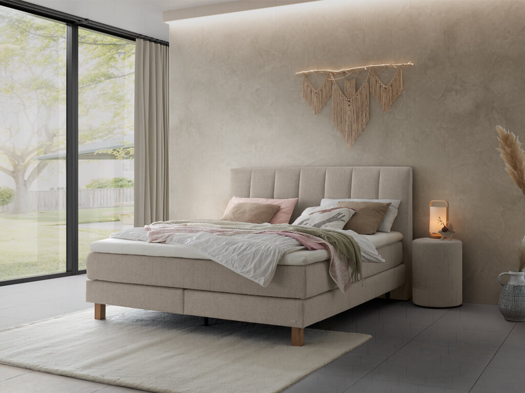 Ein Schlafzimmer, das ein hellgraues Boxspringbett mit vertikal gestepptem Kopfteil in einer ruhigen und hellen Umgebung zeigt.