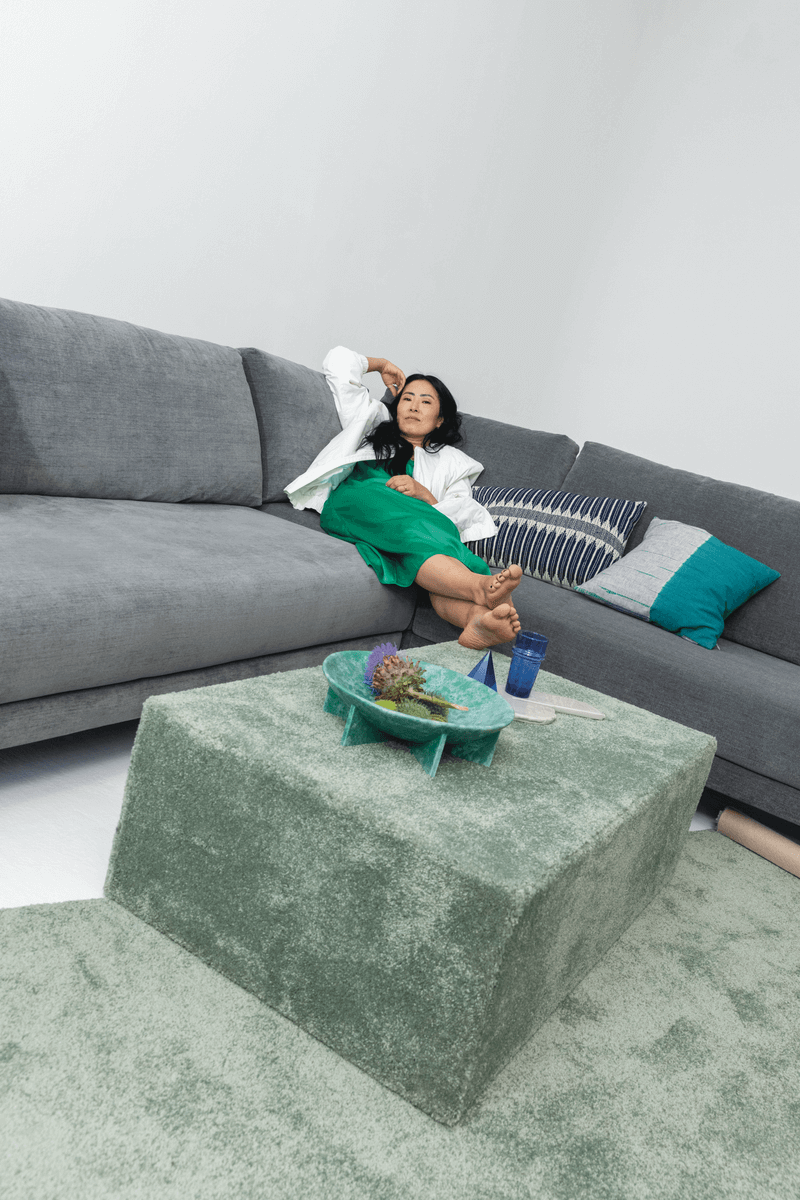 Frau liegt auf einem freistil70 Sofa in Grau. Im Vordergrund ist ein grüner Samtteppich und Couchtisch zu sehen. 