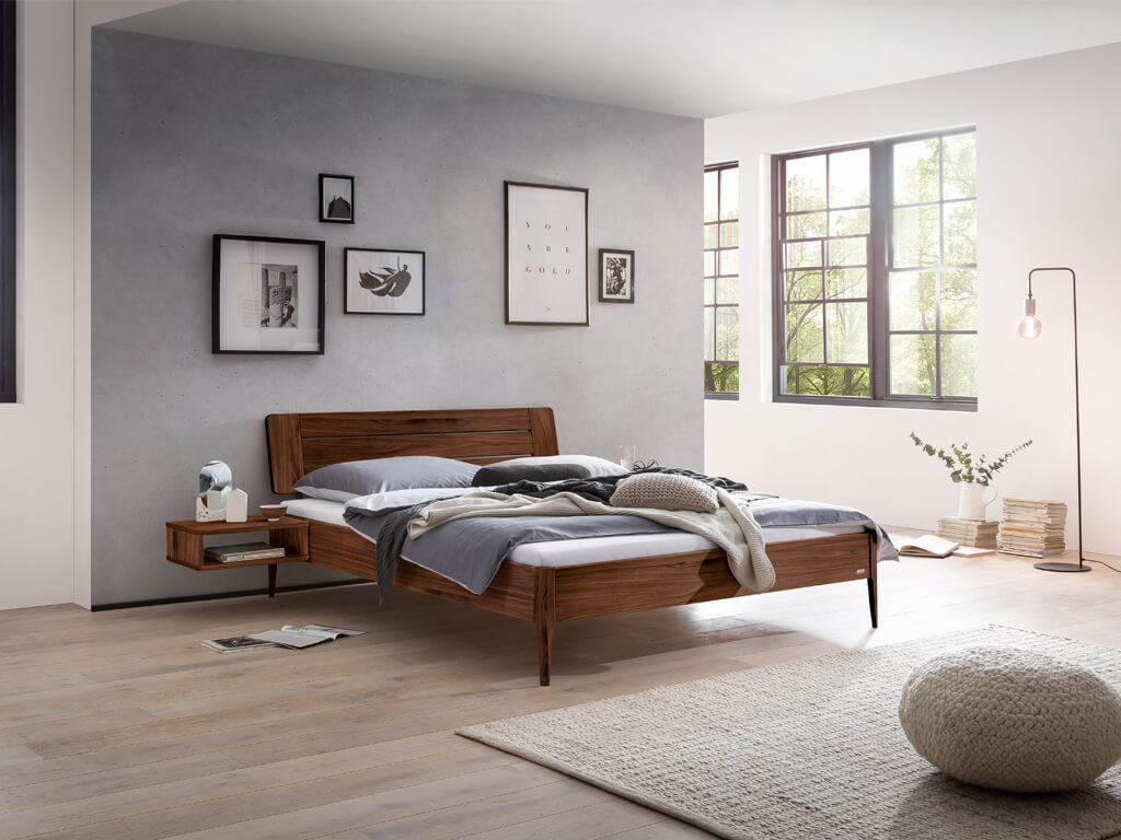 Hasena -  Lounge Bett Busseto | Holzkopfteil Kulm | Holzfuß Pile | versch. Größen & Farben konfigurierbar Eine Gesamtaufnahme eines Schlafzimmers mit dem Bettmodell "Busseto" aus Nussbaumholz. Am Bett ist ein Nachttisch in derselben Holzausführung befestigt.