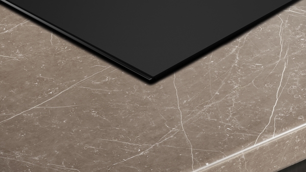 Nahaufnahme des Rands des schwarzen Induktionskochfelds, das flächenbündig in eine braune, weiß geäderte Marmor-Arbeitsplatte eingelassen ist.