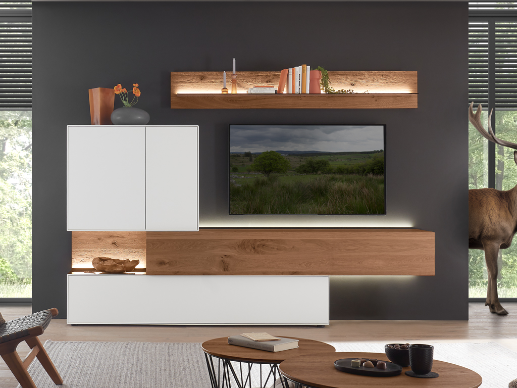 Eine moderne Wohnwand von Hartmann Torido in einem Wohnzimmer mit einem großen Fenster und Jalousien. Die Wohnwand umfasst ein schwebendes TV-Element aus Holz, einen weißen Hängeschrank und ein beleuchtetes Wandregal. 