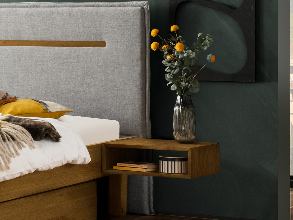 Ausschnitt eines eleganten Holzbettes mit grauem Polsterkopfteil mit Keder und Zierleiste aus Holz und Hängenachtschrank als offene Fach mit einer Blumenvase