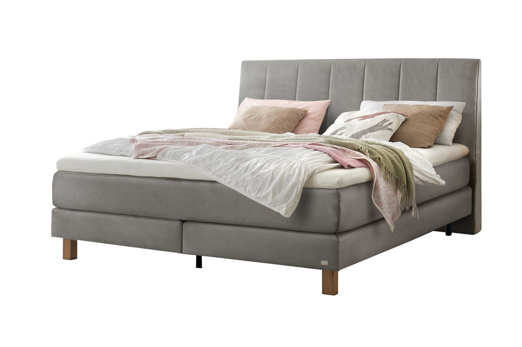 Ein Boxspringbett in einem hellen Grauton mit einem vertikal gesteppten Kopfteil, freigestellt auf weißem Hintergrund.
