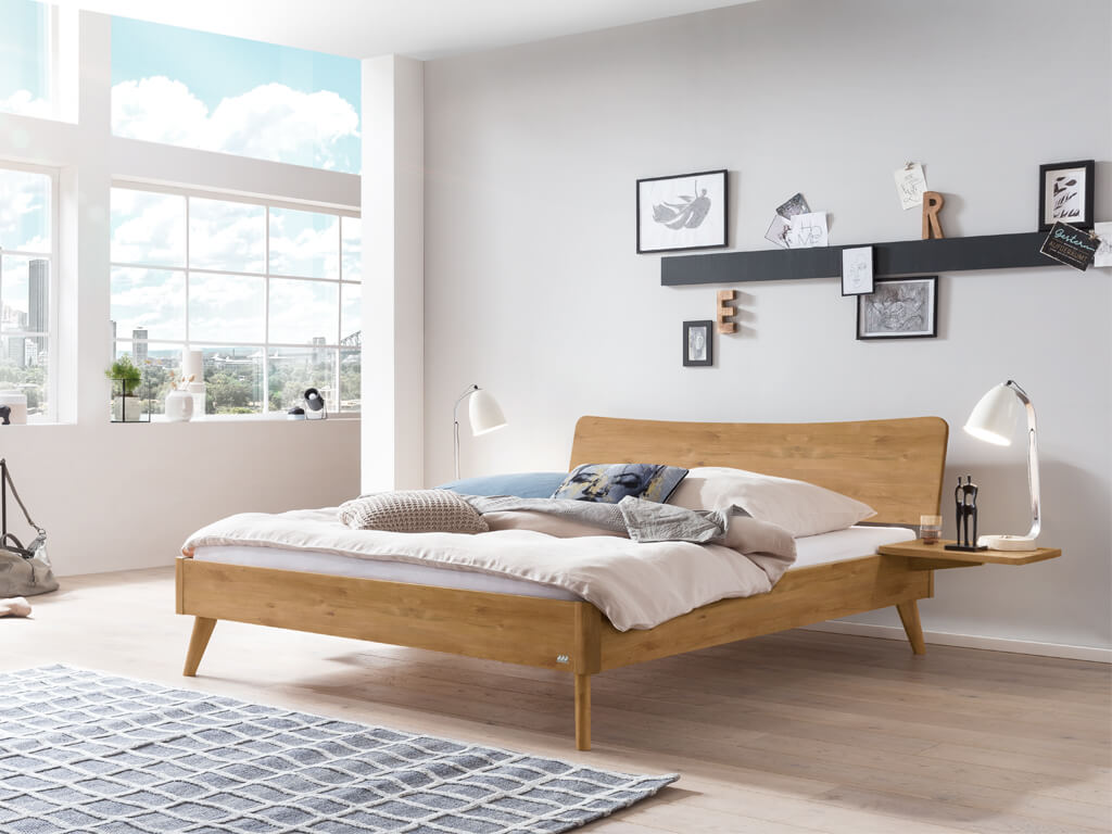 Ein Hasena Bett Caluso aus natur geölter Wildeiche in einem hellen, modern eingerichteten Schlafzimmer mit großen Fenstern. Das Bett hat ein geschwungenes Kopfteil und schräg ausgestellte Beine.