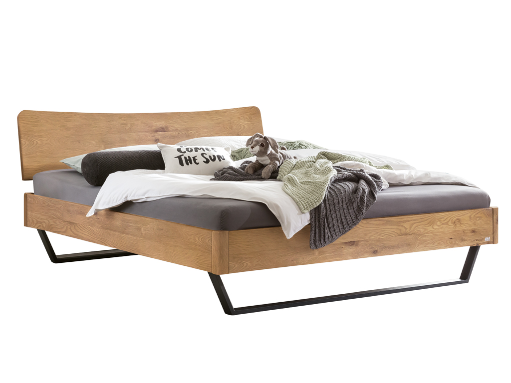 Ein Hasena Bettmodell Tresali aus natürlich geölter Wildeiche, mit einem geschwungenen Kopfteil und schwarzen Metallfüßen, das mit Bettzeug und Kissen ausgestattet ist.