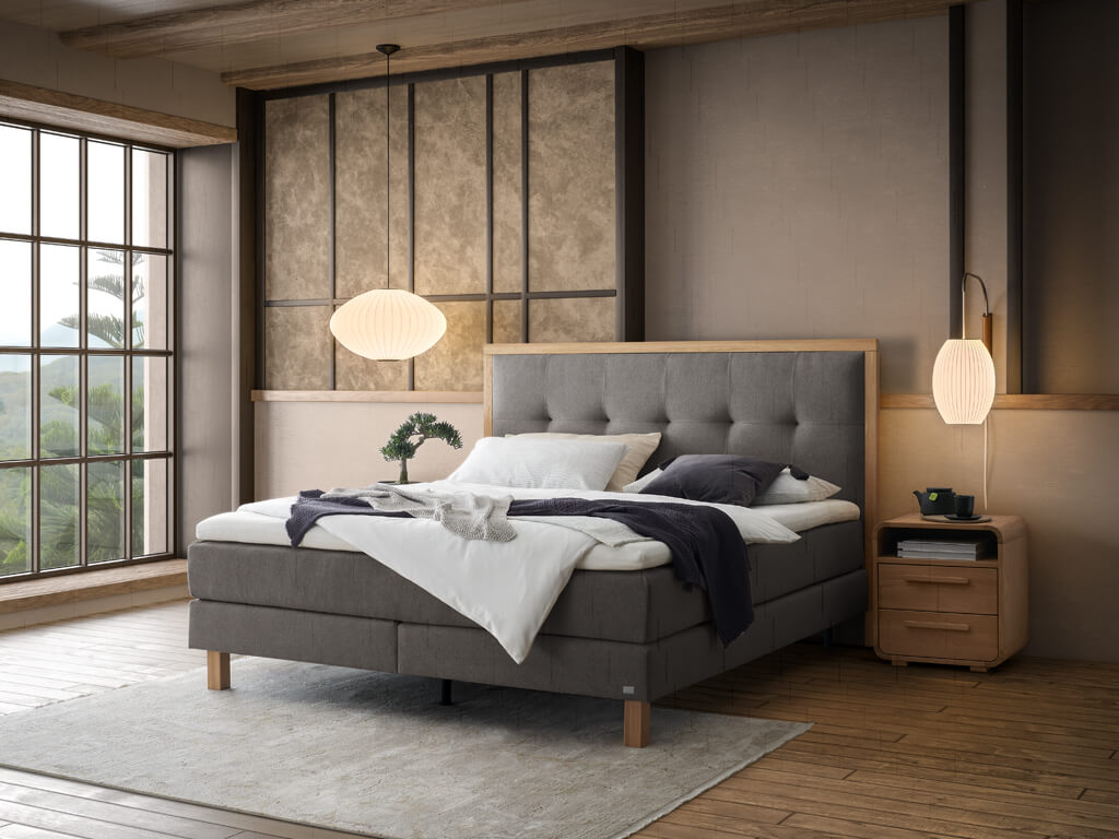 Ein Hasena Boxspringbett Pronto Solara Presley mit grauem Stoffbezug, das in einem Schlafzimmer mit einer schlichten, modernen Ästhetik steht.