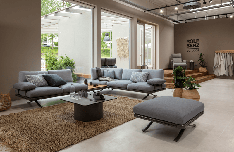 Elegantes Wohnzimmer-Set von Rolf Benz, bestehend aus einer grauen Sofagarnitur, Couchtisch und Ottoman, präsentiert in einem modernen, lichtdurchfluteten Showroom.