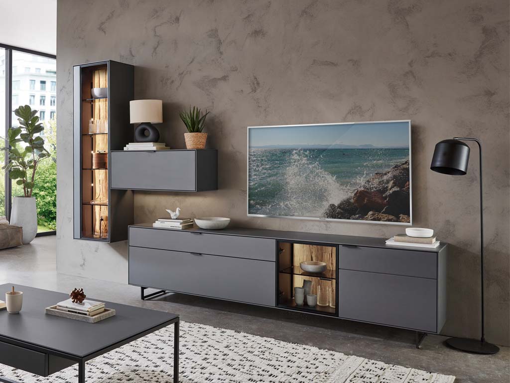 Wöstmann WM 2470 - Wohnwand  | Type 0006/0106 Wohnzimmer mit grauen Möbeln, Wandregal, Flachbildschirm und Stehlampe vor einer dekorativen Wand.