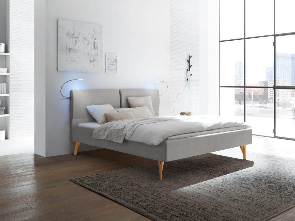 Ein graues Polsterbett mit geteiltem Kopfteil und hellen Holzfüßen in einem modernen Schlafzimmer mit viel Tageslicht.