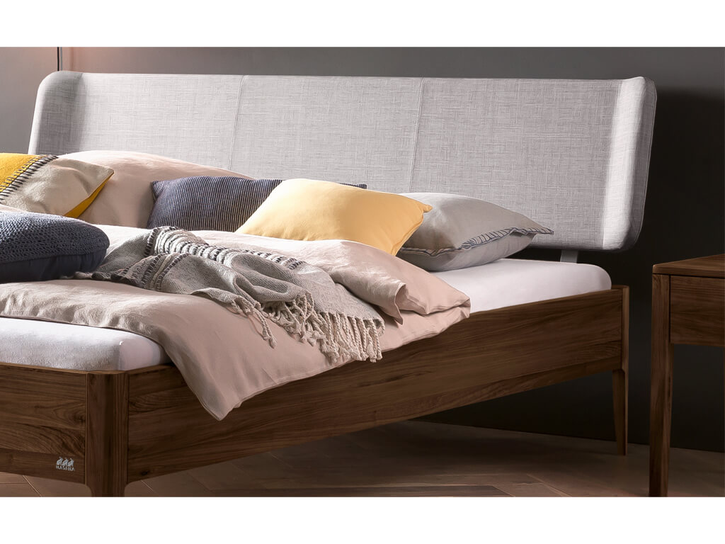Eine Nahaufnahme des oberen Teils eines Bettes Pienza in dunklem Nussbaum. Das Bett hat ein gepolstertes Kopfteil in hellem 'Pepe-grey' Stoff und ist mit verschiedenen Kissen in Beige, Grau und Gelb sowie einer gestrickten Decke dekoriert.