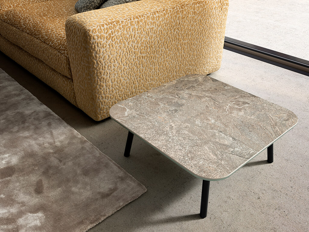 designwerk Tische - Couchtisch Four Leg | H: 29 cm Ein quadratischer Couchtisch mit Marmorplatte steht neben einem gemusterten Sofa auf einem neutralen Teppich in einem modernen Wohnzimmer.