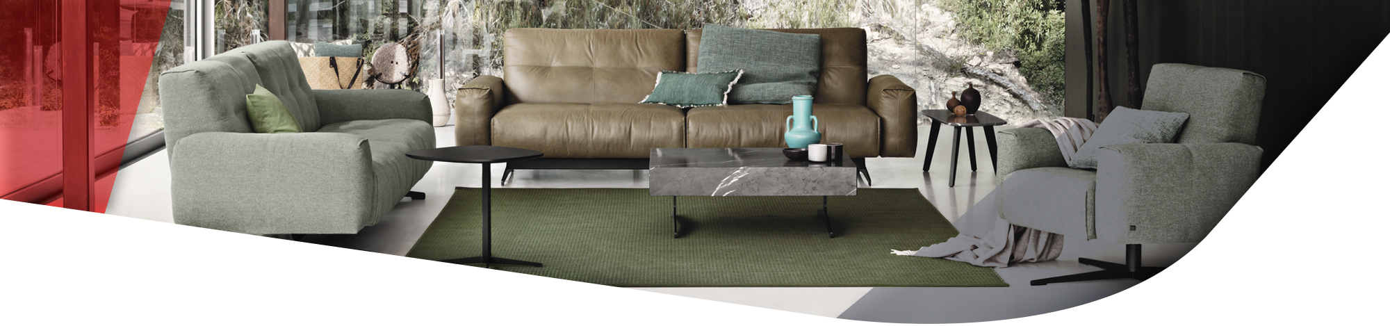 Das Bild zeigt ein modernes Wohnzimmer mit großen Glasfenstern, die einen Blick auf einen dichten Wald bieten. Der Raum ist mit einem braunen Ledersofa, zwei grünen Stoffsesseln und einem kleinen Couchtisch eingerichtet.