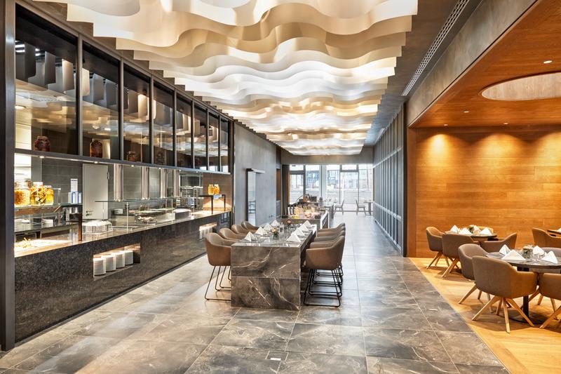 Ein elegantes, modernes Restaurant mit wellenförmiger Decke, Marmortischen und komfortablen Stühlen. Große Fenster sorgen für viel Tageslicht.
