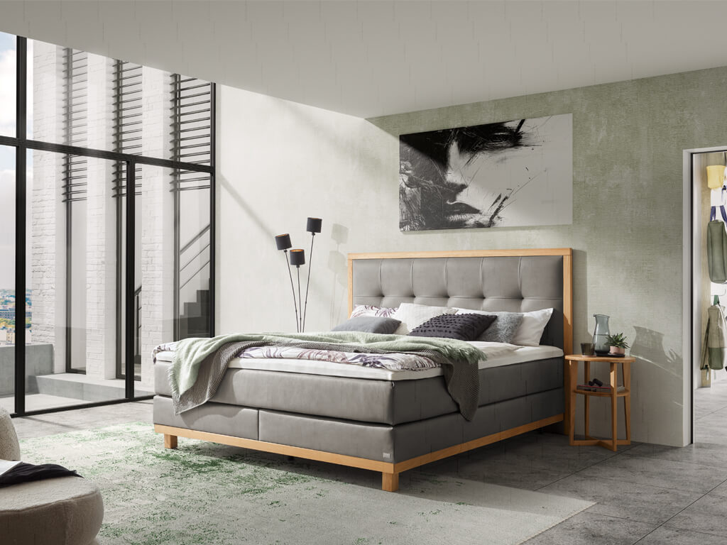 Ein modernes Schlafzimmer mit einem grauen Hasena Boxspringbett "Pronto Cecilia", das einen Stahlgrau-Bezug und einen hellen Holzrahmen hat, und einem abstrakten Kunstwerk an der Wand.