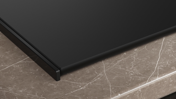 Detailansicht der Kante des Induktionskochfelds, das auf einer hellbraunen, marmorierten Arbeitsplatte aufliegt. Die Kante ist flach und hat einen dezenten, dunklen Rahmen.