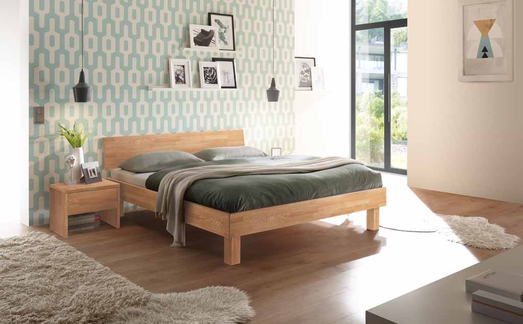 Hasena Trento - Bett Forti | Eiche bianco, geölt Komfortables Schlafzimmer mit einem Holzbett, grüner Bettwäsche, gemusterter Tapete und moderner Dekoration in einem hellen Raum