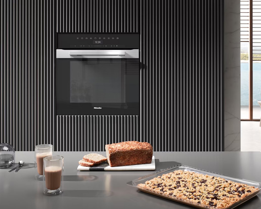 Ein moderner, schwarzer Miele Backofen, der in eine Wand mit einem vertikalen Streifenmuster eingebaut ist. Im Vordergrund steht ein Gitterblech mit einem Crumble-Kuchen und ein Laib Brot mit zwei Scheiben auf einem Brett.