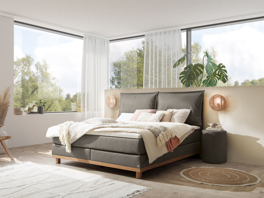 Ein Hasena Boxspringbett Pronto Zamora Dean mit stahlgrauem Stoffbezug in einem hellen, geräumigen Schlafzimmer mit großen Fenstern.