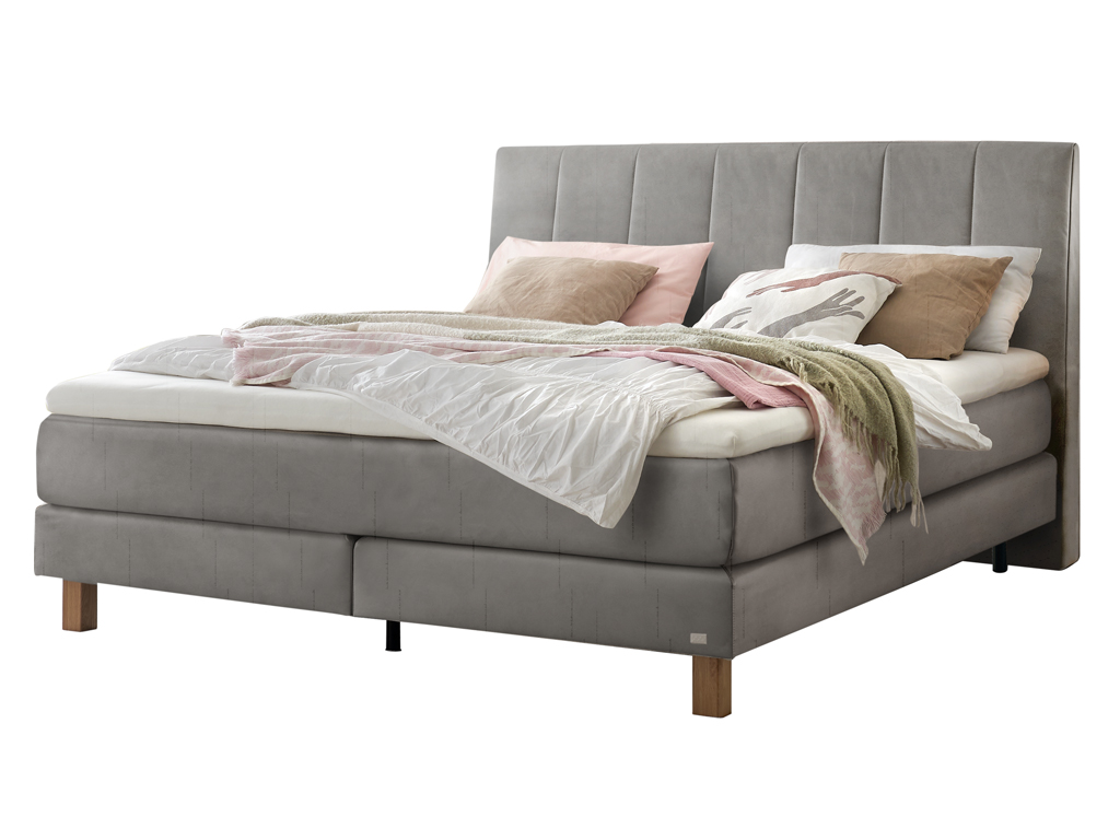 Ein Boxspringbett in einem hellen Grauton mit einem vertikal gesteppten Kopfteil, freigestellt auf weißem Hintergrund.