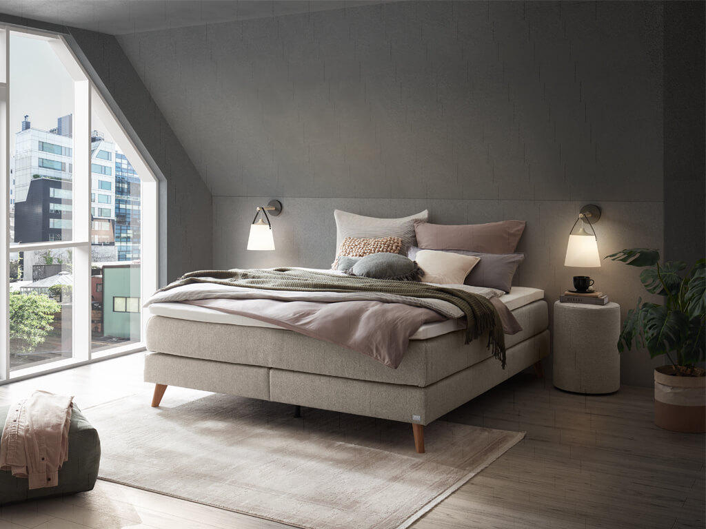Ein modernes Schlafzimmer mit einem hellgrauen Hasena Boxspringbett "Pronto Beatriz", das vor einer grauen Wand und einem großen Giebelfenster steht.