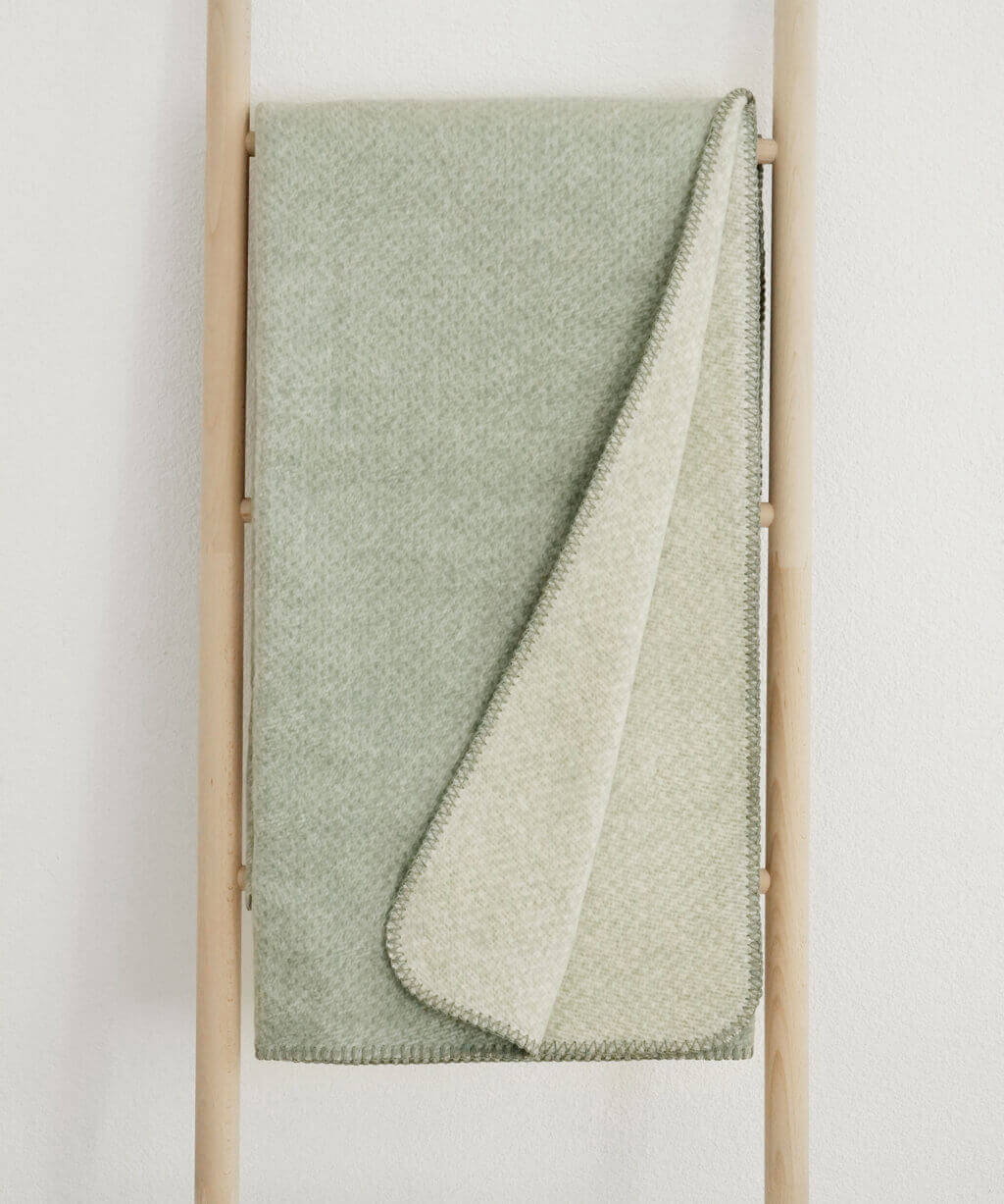 RIEMA. EMMA – Wohndecke, Bio-Baumwolle - meliert | 150 x 200 cm | grün Eine gefaltete hellgrüne Decke liegt dekorativ auf einer Holzleiter.