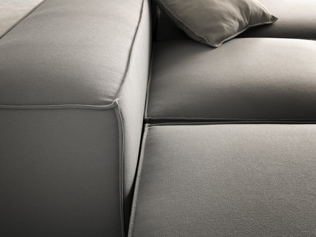 Eine Nahaufnahme des dunkelgrauen Stoffs und der Nähte eines modularen Outdoor-Sofas. Ein hellbeiges Kissen ist teilweise oben rechts im Bild sichtbar. Die Textur des Stoffes ist deutlich erkennbar.