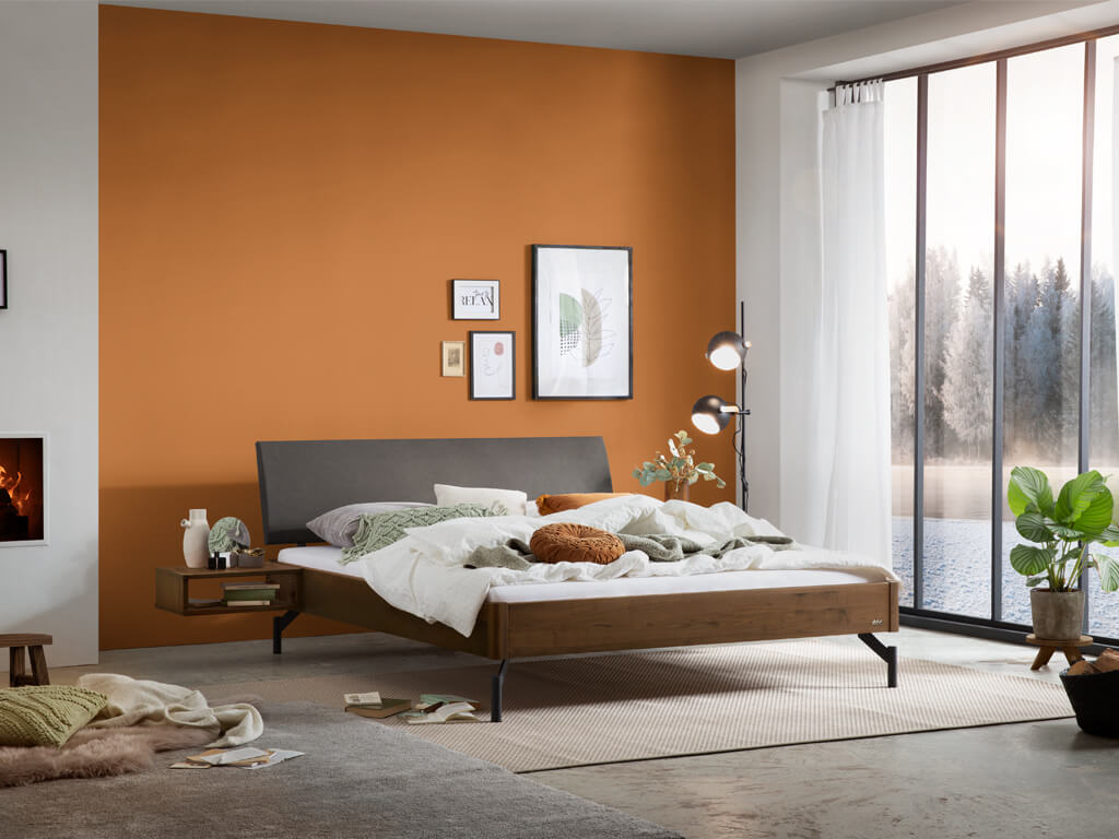 Eine Gesamtaufnahme eines Schlafzimmers mit dem Bettmodell "Broni". Das Bett hat einen Rahmen aus Nussbaumholz, ein anthrazitfarbenes Kopfteil und schwarze Metallbeine. Die Wand im Hintergrund ist in einem warmen Terrakotta-Ton gestrichen.