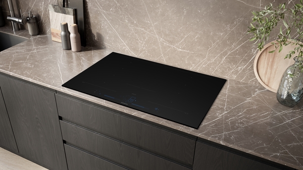 Einbauansicht des schwarzen Siemens studioLine Induktionskochfelds (EX87BNYV6E) in einer modernen Küche mit dunkelbraunen Schränken und einer graubraunen, marmorierten Arbeitsplatte.